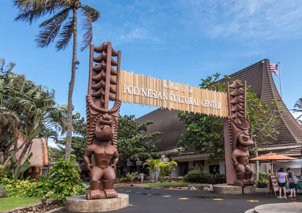 Laie, Oahu, Hawaii, USA Polynesian Cultural Center. Monumental gate into the park with brown giant aboriginal statues. People and green vegetation under blue sky.