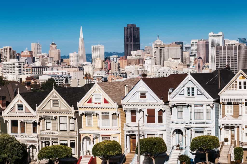 The Painted Ladies of San Francisco, California sit glowing amid the backdrop of a sunset and skyscrapers.
