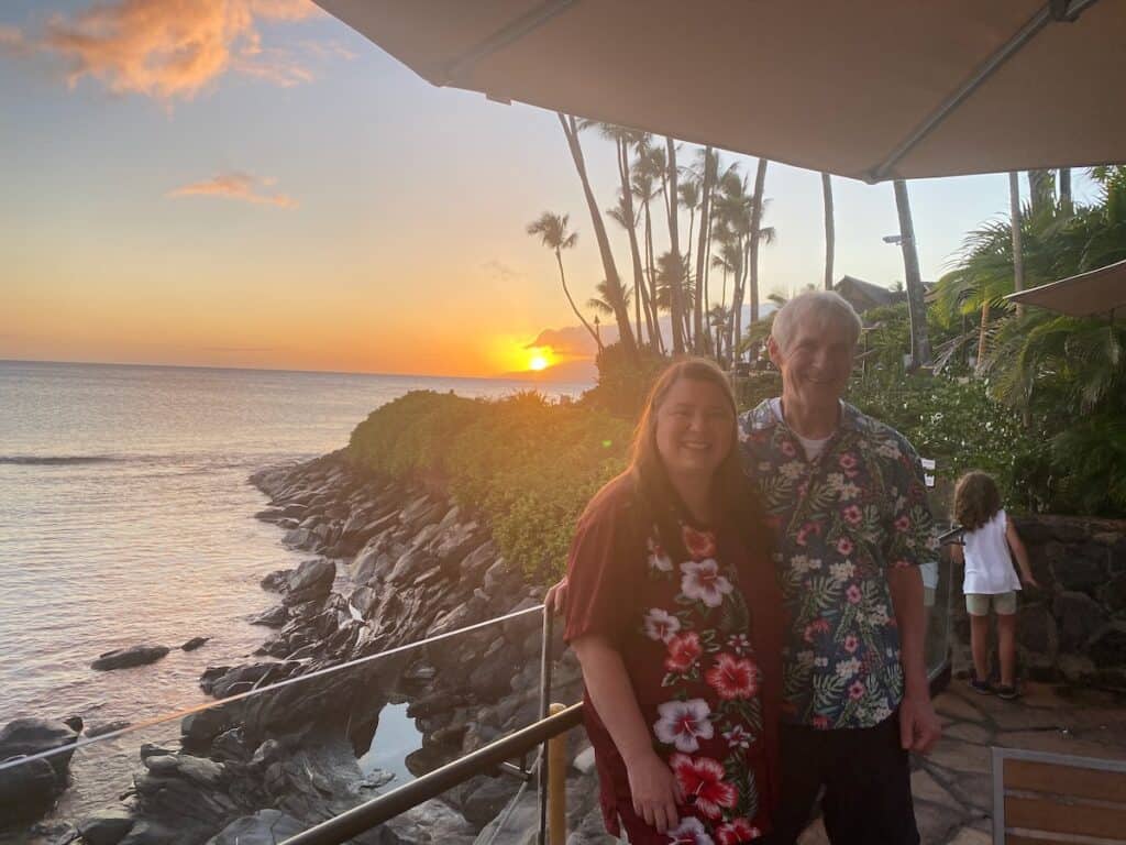 My husband and I in our Aloha shirts at Sea House standing in front of a sunset
