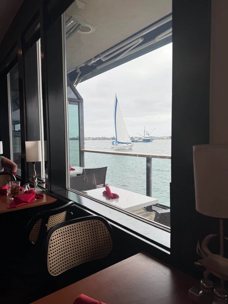 Looking through the window at the Fish Maret to an outdoor table with a sail boat passing in the background